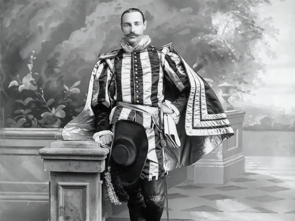  Black and white picture of John Jacob Astor IV in a period costume, leaning on a stone pillar with his top hat on his hip, wearing a diamond ring and cufflinks.